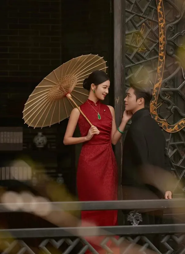 Bride in a red qipao holding a paper parasol beside the groom at a carved timber gateway