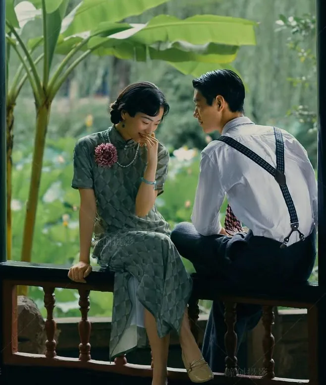 Couple in 1930s-style dress laughing together on a garden pavilion ledge beside a lotus pond