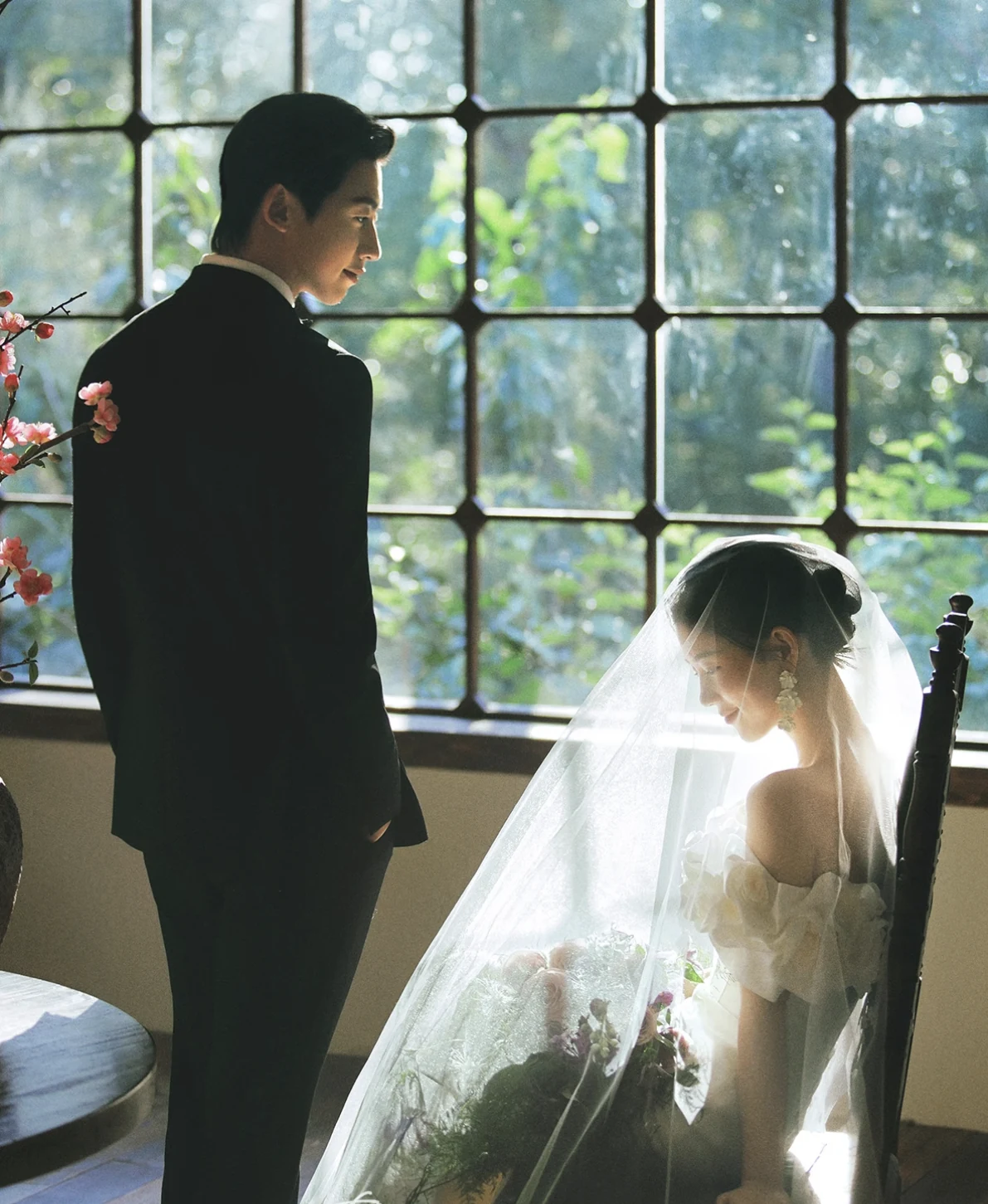 Bride in a veil seated by a tall window as the groom stands beside her in a black tuxedo